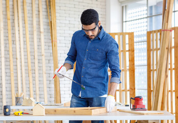 Indian Asian professional engineer architect foreman labor worker wears safety goggles glasses and gloves using square angle degree ruler measuring wood on working table in housing construction site