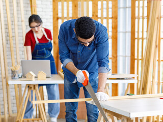 Obraz premium Indian Asian professional male engineer architect foreman labor worker wears safety goggles glasses and gloves using hand saw cutting wood plank on wooden table while female colleague working behind
