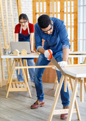 Indian Asian professional male engineer architect foreman labor worker wears safety goggles glasses and gloves using hand saw cutting wood plank on wooden table while female colleague working behind