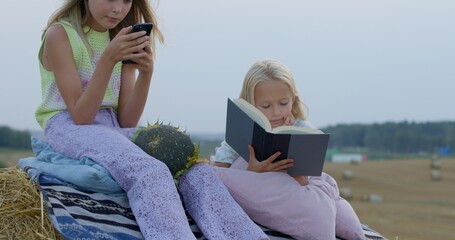 two girls eat sunflower seeds on mown rye in field. Landscape of straw bales against setting sun on background. Sisters watch video on smartphone and read book outdoors. Childhood. Country life