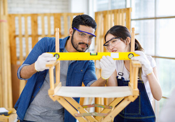 Indian professional bearded male engineer architect foreman labor worker carpenter helping Asian female colleague using water level magnetic ruler tool checking alignment while assemble wooden chair