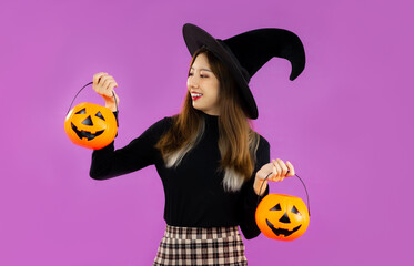 Halloween, young asian girl in black costume wearing black witch hat holding and carrying orange pumpkin bucket on purple background.