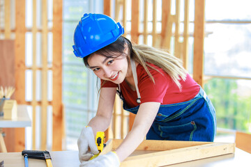 Asian professional female engineer architect foreman labor worker wears safety hard helmet jeans apron and gloves standing using squard angle degree ruler and pencil measuring wood plank on workbench