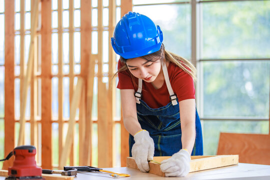 Asian professional female engineer architect foreman labor worker wears safety hard helmet jeans apron and gloves standing using squard angle degree ruler and pencil measuring wood plank on workbench