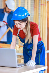 Asian professional female engineer architect foreman labor worker wears safety goggles and jeans apron standing smiling using laptop computer working on wooden table with tools in construction site