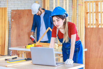 Asian professional female engineer architect foreman labor worker wears safety goggles and jeans apron standing smiling using laptop computer working on wooden table with tools in construction site