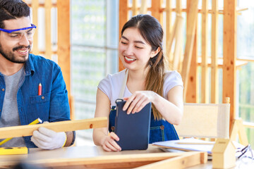 Asian professional cheerful female engineer architect foreman labor worker sitting smiling holding tablet computer discussing with Indian bearded male carpenter in workshop housing construction site