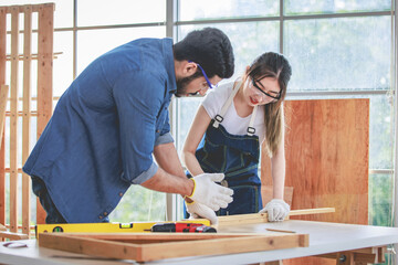 Asian young female engineer architect foreman labor worker wears safety goggles gloves and apron holdind using steel hammer pounding nail on wood stick when Indian professional male carpenter helping