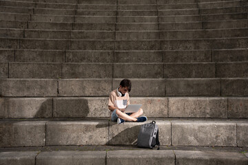 Fototapeta na wymiar Teenager boy working on laptop sitting on staircase with dramatic lighting