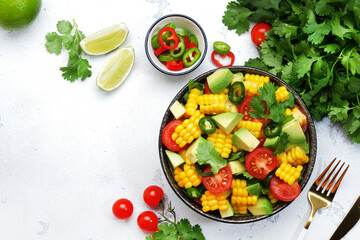 Fototapeta na wymiar Mexican spicy salad with corn, avocado, jalapeno peppers, cherry tomatoes and cilantro. White kitchen table background, top view