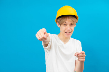 Happy handsome teen builder in a yellow helmet is looking at the camera and points fingers of both hands at you, standing in a studio on a blue background