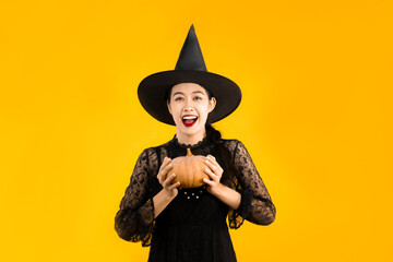 Halloween theme, happy young asian woman in witch costume black dress wearing hat holding pumpkin on yellow color background.