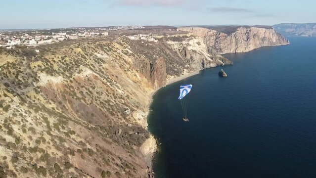Aerial drone view of a man flying a white and blue paraglider over a hill and trees to the sea waves near the rocks. Active paraglider flight over the seascape with clear skies at suset. Extreme sport