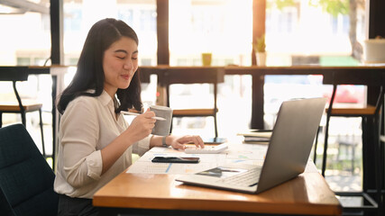 Cheerful woman office worker drinking hot coffee and using laptop computer at bright modern corporate office