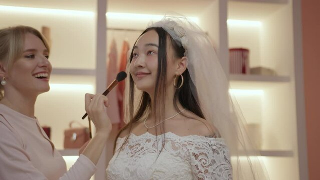 Bridesmaid applying makeup for Asian newlywed preparing for wedding ceremony standing in dressing room.