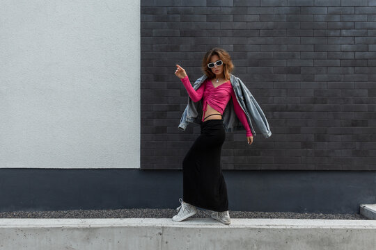 Stylish beautiful trendy hipster redheaded woman with cool white sunglasses wearing trendy bright clothes with fashion denim jacket, pink top, skirt and sneakers dancing near the wall in the street