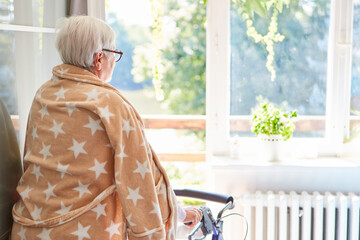 Alte Frau im Bademantel steht mit Rollator am Fenster