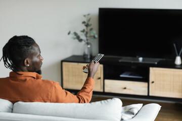African american man watching television, pointing remote control at empty TV screen, mockup