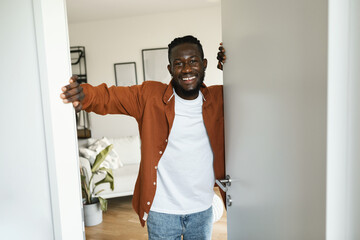 Happy african american man opening door of his new apartment and smiling at camera, standing indoors