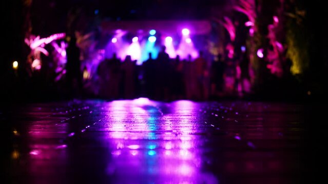 Bright stage lighting and dark people figures, blurred view of music show or performance. Dark alley, camera focused on foreground, light reflection at wet pavement
