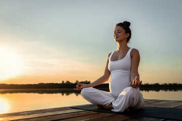 Naklejka na ściany i meble Pregnant woman doing yoga at lake during sunset.