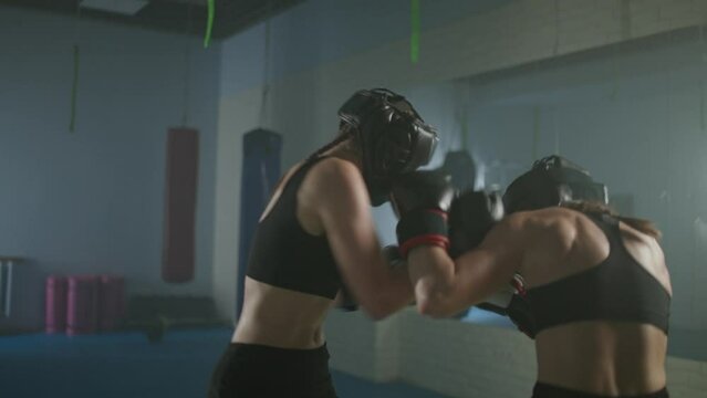 Combat sparring of two females fighters wearing protective helmets, training in the boxing gym, females train defence and series of punches on a battle, motion blur.