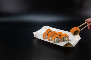 A cardboard box for sushi, a man hand using chopsticks takes a roll. Black background. Place for...