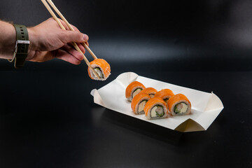 A cardboard box for sushi, a man hand using chopsticks takes a roll. Black background. Place for...