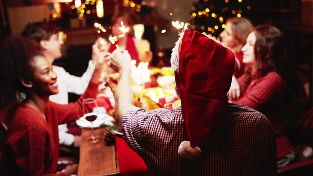 Groups of people celebrate Christmas eve while playing sparkler and have dinner at night which have many food and wine on the table.