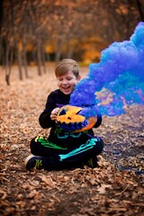 Very smiling Young European boy seven years old dressed the colourful Jack Skellington costume, holding a big pumpkin with the blue smoke in front of him in the autumn yellow park on the Halloween