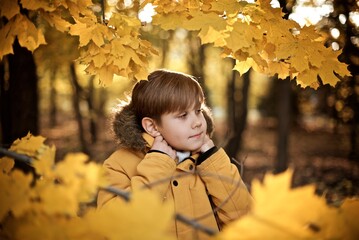 Very smiling boy seven years old in the yellow jacket is standing in the autumn park behind the...