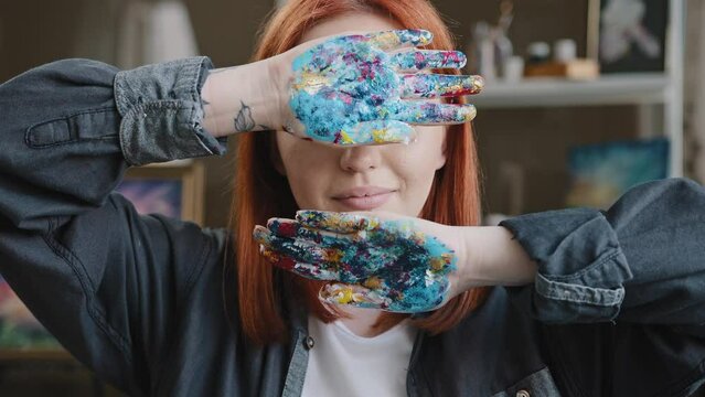 Close up hipster student art class lady Caucasian red-haired girl artist woman painter designer showing dirty palms in paints holding hands covers face with arms after drawing looking at camera indoor