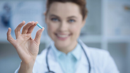 blurred and cheerful doctor in white coat smiling while holding capsule.