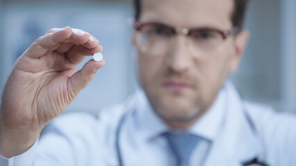 blurred doctor in glasses and white coat looking at camera and holding pill.