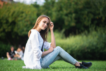 Naklejka na ściany i meble Portrait of a happy Caucasian girl in casual clothes who is sitting on the green grass in nature in the park.