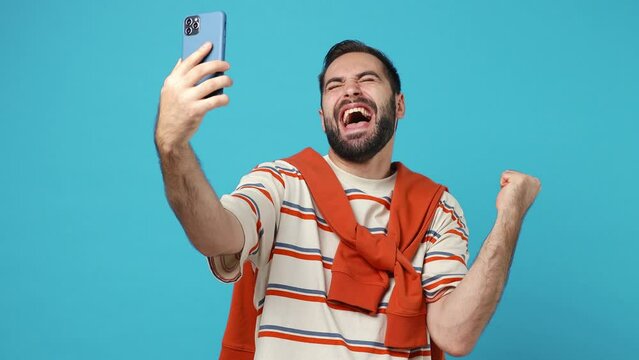 Overjoyed young brunet man 20s years old wears striped t-shirt doing selfie shot on mobile phone post photo on social network doing winner gesture isolated on plain pastel light blue background studio