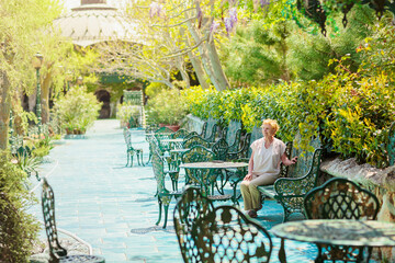 Mature attractive woman traveler sitting alone on the terrace of coffee shop in beautiful park in Bulgaria. Active life of the elderly in retirement, active seniors