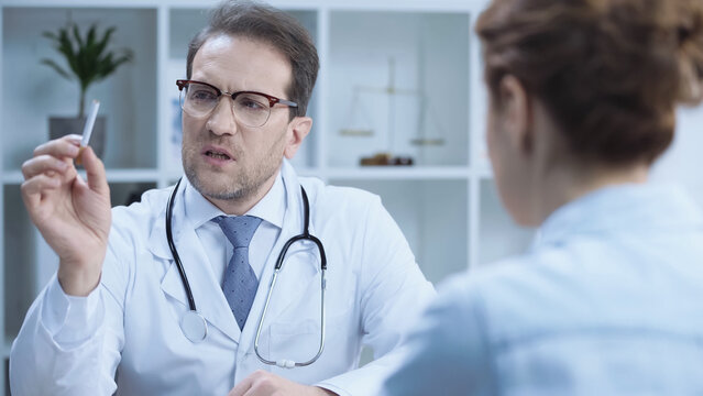 Serious Doctor In Glasses Holding Cigarette While Warning Blurred Patient In Hospital.