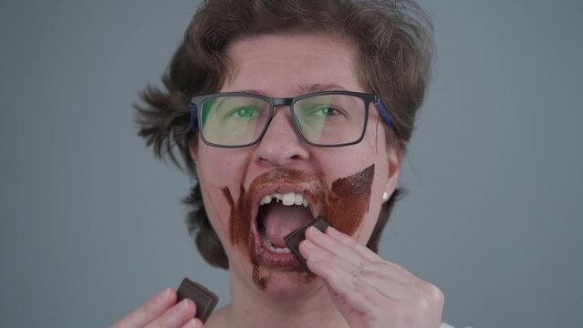 A adult woman with missing teeth is smearing chocolate on her face in the studio. The topic of diabetes, the dangers of sugar, teeth and candy. Diet and healthy eating. A funny female all in chocolate