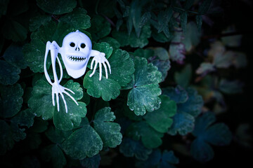 White plastic figurine skull-ghost with bones in the green leaves background at night. Copy space