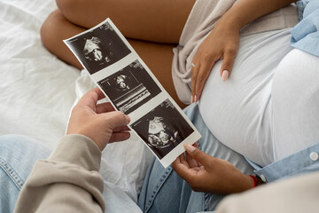 Multiethnic pregnant couple watching ultrasound image picture at home. Closeup feeling happy, stroking tummy and excited on seeing baby ultrasound
