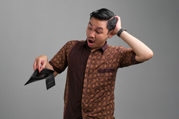 Shocked young Asian man wearing batik shirt looking into empty wallet while touching head isolated...