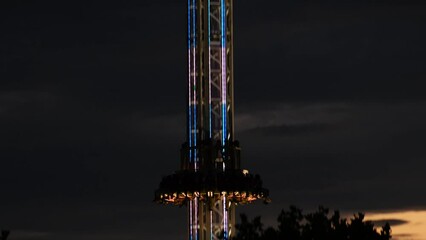 Big drop ride at night. Close-up of a free fall or high gravity or drop tower with colourful neon lights against the dark blue sky. Carnival, amusement or thrill or theme park, entertainment and fun.