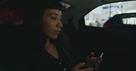 Woman using cellphone in car backseat at night. Person riding taxi cab in urban city street typing message on smartphone device commuting from work