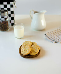 Cookies on a wooden plate on a white table. A jar of coffee and tea, a glass of milk, a white jug, a napkin.