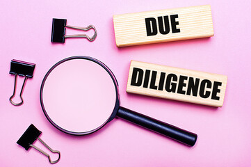 On a pink background, a magnifier, black paper clips and wooden blocks with the text DUE DILIGENCE....