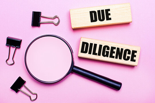 On a pink background, a magnifier, black paper clips and wooden blocks with the text DUE DILIGENCE. Business concept