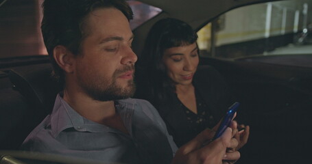 Young man and woman in car backseat talking and smiling. Male colleague showing smartphone device screen to friend in cab transportation. People in commute after work