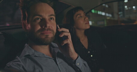 People riding taxi cab in car backseat at night. Young man and woman passengers in the evening after work talking on phone