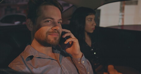 People riding taxi cab in car backseat at night. Young man and woman passengers in the evening after work talking on phone